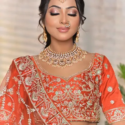 A classic bridal portrait featuring a beautiful Kundan choker with pearl details. This set is paired with a matching maang tikka and a delicate nath, complementing the warm tones of the orange lehenga.