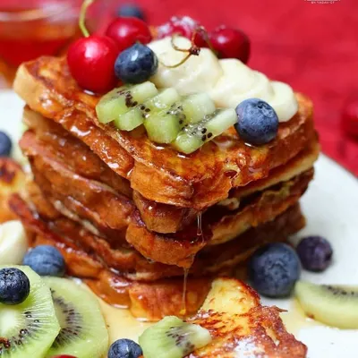A tall stack of golden French toast is topped with fresh berries, kiwi, and bananas, with syrup dripping down the sides. This close-up food photograph is vibrant, delicious, and expertly styled.