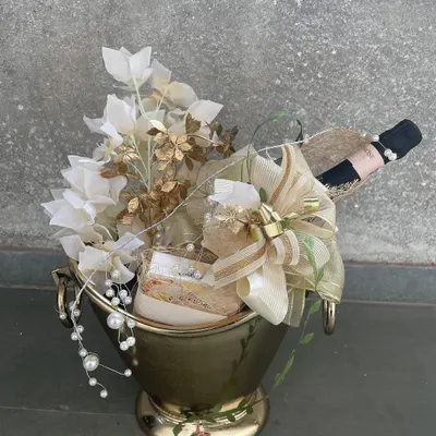 The epitome of luxury bottle gifting. A bottle of champagne is nestled within a golden bucket, lavishly decorated with white and gold florals and cascading pearls, creating a truly show-stopping gift.