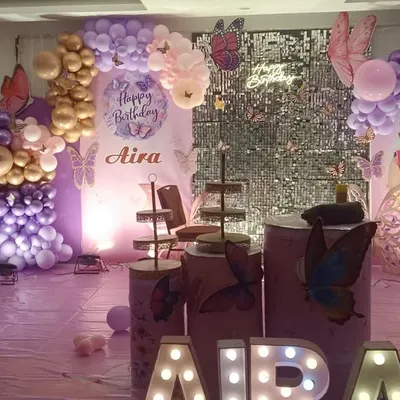 A beautiful butterfly theme for a first birthday, featuring shades of purple and pink. The decoration includes a silver shimmer wall, personalized backdrops with the child's photo, and marquee letters spelling her name.