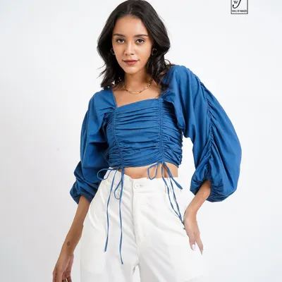 A crisp studio shot for a lookbook, featuring a model in a stylish blue ruched top and white trousers. The clean background ensures the clothing is the main focus.