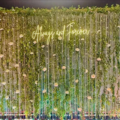 A romantic photo backdrop with a "Always and Forever" neon sign. The wall of cascading greenery, fairy lights, and scattered white roses creates an enchanting, garden-like effect.