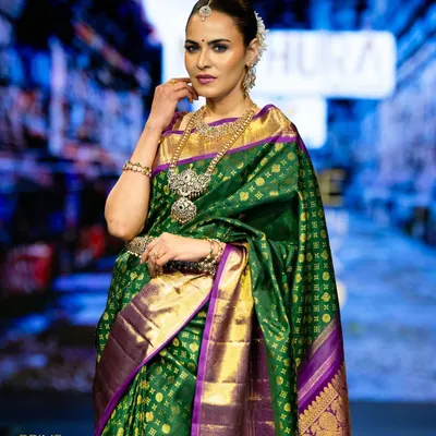 A model graces the runway in a stunning green and purple Kanchipuram silk saree, its rich gold border catching the light with every step.