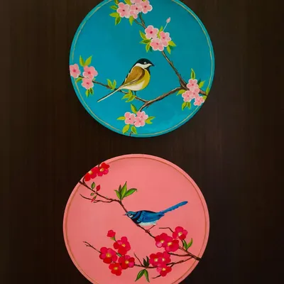 A beautiful pair of nature-themed wall plates. One features a bird on a cherry blossom branch against a teal background, and the other a blue bird on a pink background, creating a perfect complementary set.