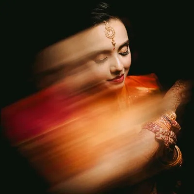 An ethereal bridal portrait created with a slow shutter speed. The motion blur of her orange saree creates a beautiful, painterly effect, while her serene expression remains in focus, blending art with tradition.