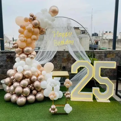 A 25th birthday celebration on a rooftop terrace. This setup features a semi-circular balloon arch in rose gold, peach, and white, a sheer canopy, a neon sign, and large marquee numbers.