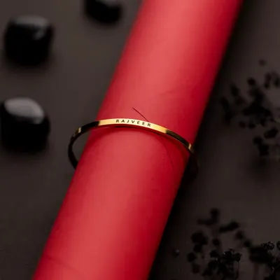 This minimalist shot places a custom-engraved gold bracelet on a bold red cylinder. The contrast in color and texture makes the delicate bracelet pop, while the scattered dark stones add a touch of elegance.