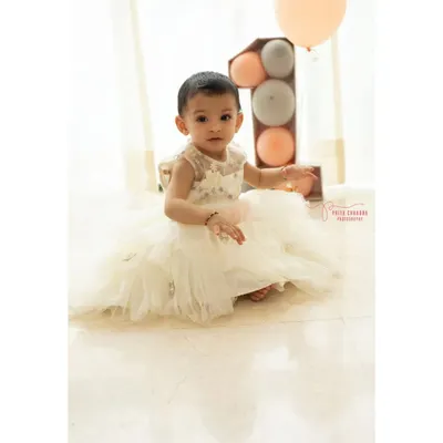 A little princess in her beautiful white dress, celebrating her very first birthday. This minimalist studio setup with soft, natural light and pastel balloons puts all the focus on her sweet and curious expression.