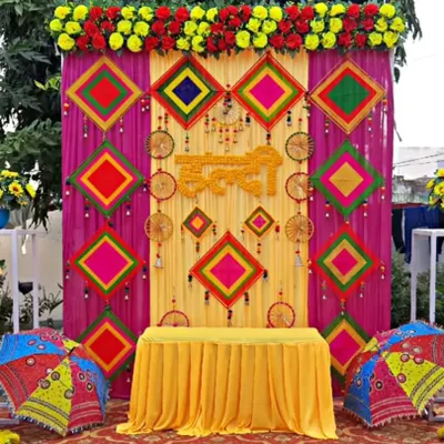 A truly grand Haldi backdrop featuring a vibrant mix of yellow and magenta drapes, decorated with colorful threadwork panels and dreamcatchers. The setup is framed with marigold garlands and Rajasthani-style umbrellas for a festive, traditional look.
