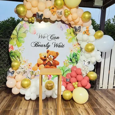 A charming teddy bear themed baby shower decoration with the message "We Can Bearly Wait". The setup features a custom circular backdrop framed by a balloon arch in pastel yellow, peach, and gold.