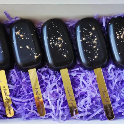 A set of sleek and modern black cakesicles, each one speckled with edible gold for a touch of luxury. They are presented on custom gold sticks that read "a treat of goodness".