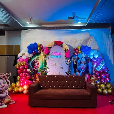 A full view of our grand Krishna themed baby shower stage. The setup includes two large character backdrops, a central panel with a blessing, a neon 'Baby Shower' sign, and a comfortable sofa for the parents to be, all set on a red carpeted stage.