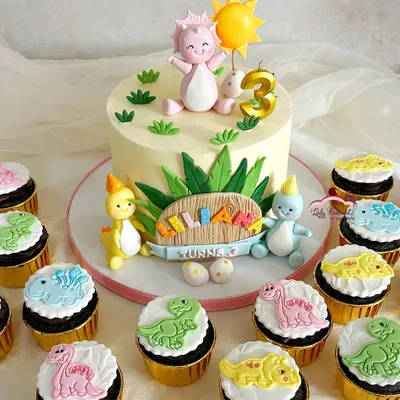 This friendly dinosaur cake was the centerpiece of a 'Dino-mite' third birthday. The main cake is surrounded by matching cupcakes, each featuring a different colorful dinosaur, creating a full and adorable dessert display.