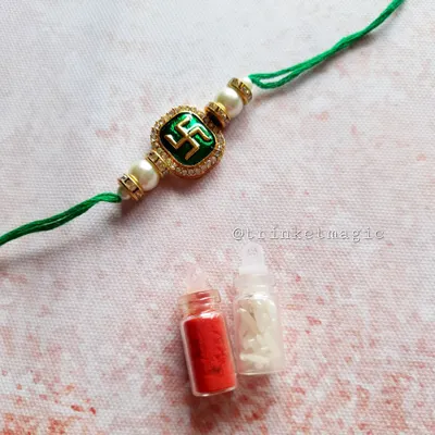 A simple and sacred Swastik Rakhi. This design features a green enamel Swastik symbol with pearl details on a traditional green thread, and it comes with a small bottle of Roli and Chawal.