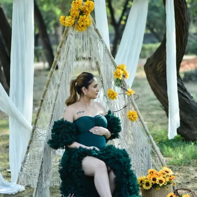 A beautiful boho-themed outdoor maternity shoot. The mom-to-be is seated in a macrame teepee adorned with yellow flowers, wearing a stunning dark green ruffled gown from my collection.