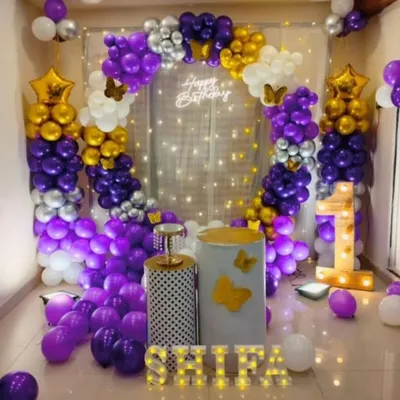 A rich purple, gold, and white theme with butterfly details. This setup includes a full balloon circle against a fairy light curtain, a light-up number '1', and marquee letters for the name 'SHILPA'.