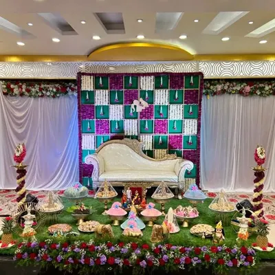 A full view of a grand Seemantham stage. The decor features a checkered floral backdrop in purple and white, a classic white chaise lounge, and an extensive display of decorated plates on an artificial grass carpet.
