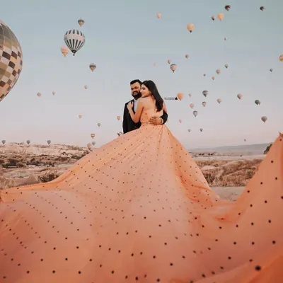A breathtaking wide-angle shot showing the full expanse of the peach polka-dot gown's trail against the sky full of hot air balloons in Cappadocia. This is how you create a truly epic pre-wedding photo.