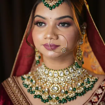 A classic North Indian bridal look featuring a deep red lehenga and stunning kundan jewelry with green accents. The makeup is focused on defined, smokey eyes and a bold lip to create a timeless and royal feel.