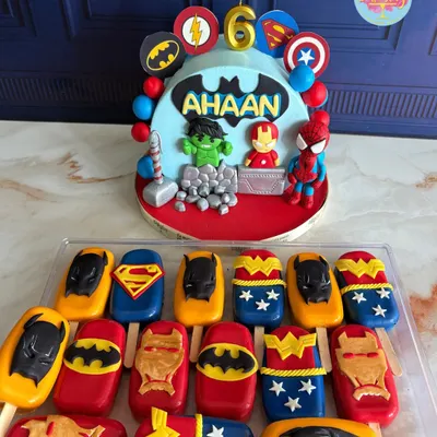 A superhero-themed dessert spread for a 6th birthday party. The main cake features fondant figures of Hulk, Iron Man, and Spider-Man, complemented by matching cakesicles with logos of Batman, Superman, and Wonder Woman.