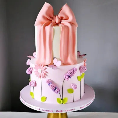 This elegant two-tiered cake is a perfect example of the popular "bow cake" trend. It features a tall top tier wrapped in a dramatic, handcrafted pink fondant bow, with the bottom tier decorated with delicate, piped wildflowers.