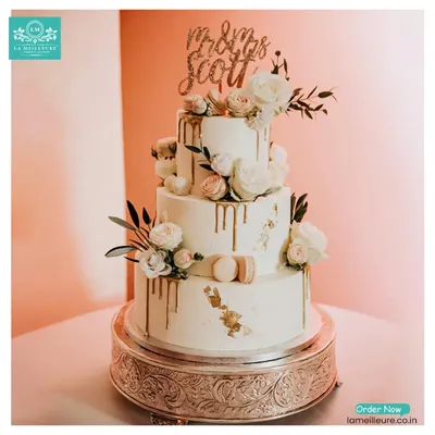 A stunning three-tiered custom wedding cake. This elegant creation features a white buttercream finish, a delicate gold drip, and is adorned with fresh flowers and macarons.