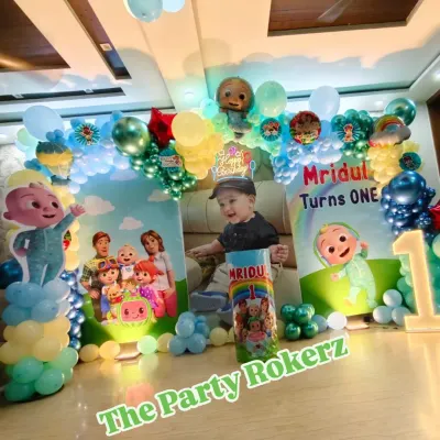 A complete Cocomelon-themed setup for a first birthday at home. It includes a balloon arch, a large backdrop with a photo of the birthday boy, character cutouts, and a light-up number.