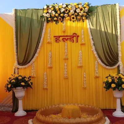 A classic Haldi ceremony backdrop with a bright yellow and green color scheme. The top floral arrangement uses fresh sunflowers and white flowers, and the 'Haldi' sign is made from red floral letters.