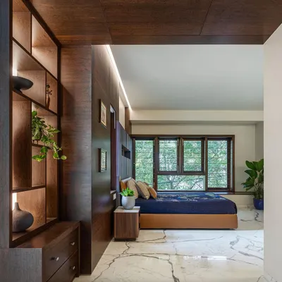 This bedroom in the Kanasu project is designed as a tranquil retreat. A custom wood-paneled wall incorporates a display niche and side table, while large windows frame the view of the trees outside.