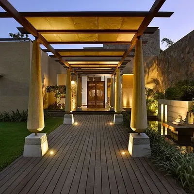 The entrance to C Farm in Chattarpur is marked by a wooden deck and a pergola supported by unique, cone-shaped columns. This walkway is flanked by a stone wall and a water channel, creating a multi-sensory journey into the home.