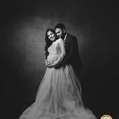A dramatic and romantic black and white maternity portrait. The use of a dark, textured backdrop and a flowing gown creates a fine art feel, focusing on the couple's elegant pose and connection.