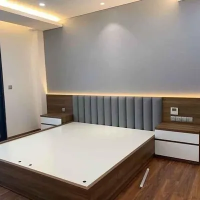 A modern bed with an extended wooden headboard and integrated side tables. The headboard features a comfortable grey upholstered panel and warm, ambient backlighting, creating a cozy and stylish focal point.