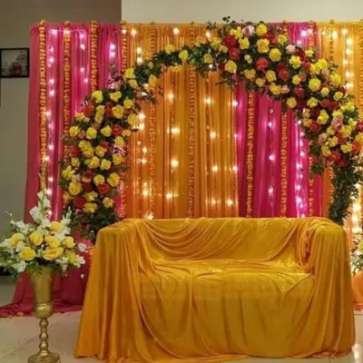 For pre-wedding functions like Haldi or Mehendi, I create vibrant backdrops that are perfect for photos. This setup features a beautiful arch of fresh yellow and red roses against rich pink and orange drapes, with fairy lights to add a warm glow.