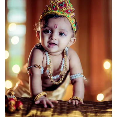This portrait captures the bright, curious expression of a baby dressed as Lord Krishna. The warm, glowing lights in the background create a beautiful bokeh effect, and the detailed costume, from the peacock feather crown to the pearl necklaces, is arranged perfectly.