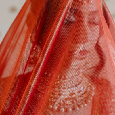 A bride's serene expression seen through her sheer red dupatta, a moment of quiet contemplation.