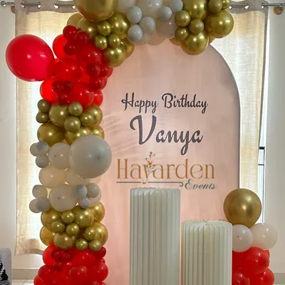 A simple yet elegant birthday setup at home. The arched backdrop with a custom name and a red, gold, and white balloon garland creates a beautiful focal point.