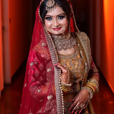 A classic bridal portrait showing the bride in her stunning red and gold lehenga. The warm lighting highlights her intricate jewelry and confident smile.