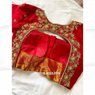 A stunning red bridal blouse with intricate floral embroidery in contrasting green and gold. The deep back cut is designed to be elegant and is perfect for pairing with a traditional silk saree.