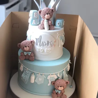 The cutest two-tiered "Mom to Be" cake for a baby shower. This design in pastel blue and white features adorable teddy bears, baby clothes, and a crown topper.