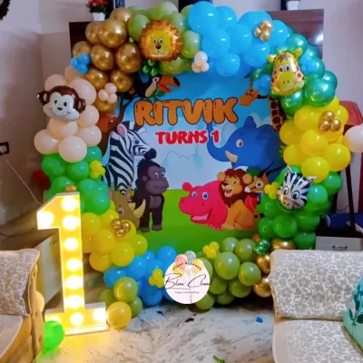 A fun jungle safari theme for a first birthday party at home. This setup includes a custom backdrop with cartoon animals, a colorful balloon arch, and a light-up number '1'.