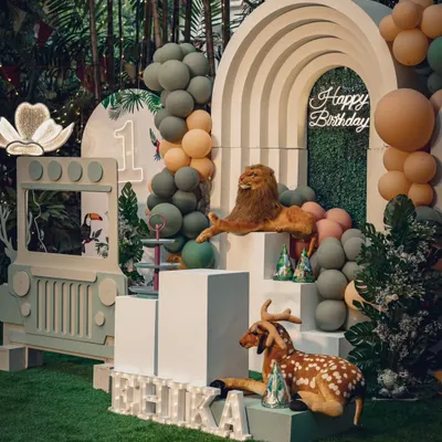A sophisticated safari theme for a first birthday. This setup uses a beautiful rainbow arch, a green hedge backdrop, and realistic animal props to create a chic and wild celebration.