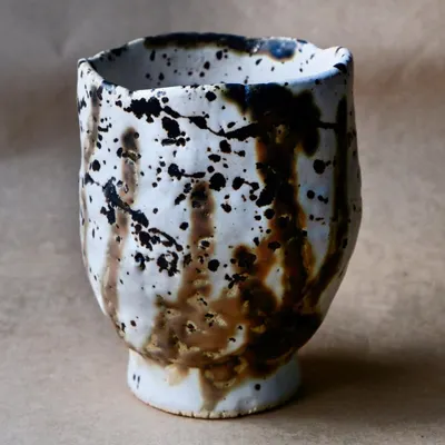 This handmade ceramic cup features a dynamic splash of black and brown glaze over a white base. The irregular rim adds to its unique, handcrafted character.
