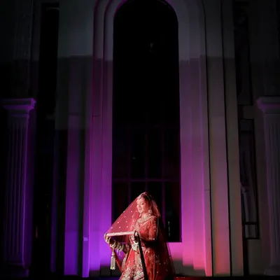 A dramatic bridal portrait using purple uplighting and architectural elements. The bride, in her red lehenga, looks regal and mysterious, like a queen in her palace.
