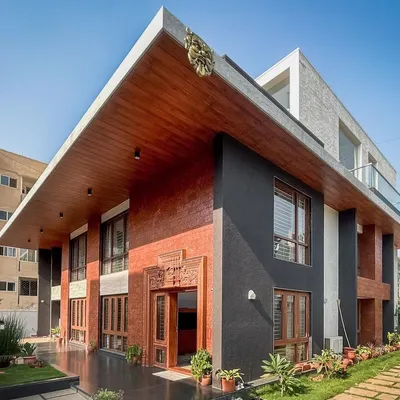 The facade of a contemporary villa in Hennur, showcasing a dynamic composition of exposed brick, dark textured surfaces, and a prominent cantilevered roof.