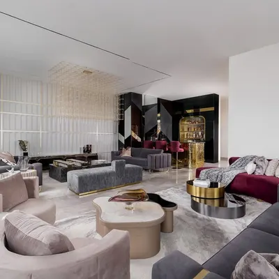 An expansive view of a complete entertainment lounge we designed for hosting and relaxation. This space features multiple seating zones with custom curved and channel-tufted sofas, a sculptural grid chandelier, and a dedicated bar area, all tied together with a palette of greys, blush, and rich burgundy.
