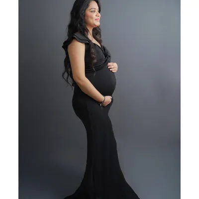 A timeless and elegant solo maternity portrait. This side profile shot beautifully showcases the baby bump, with the mother-to-be in a classic black gown against a solid grey studio background.