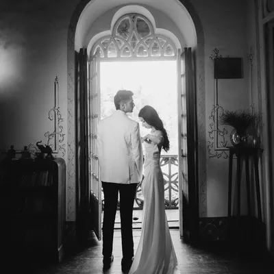 A dramatic, cinematic portrait of a couple standing in a grand doorway. The high contrast black and white emphasizes their elegant silhouettes and the promise of the moment.