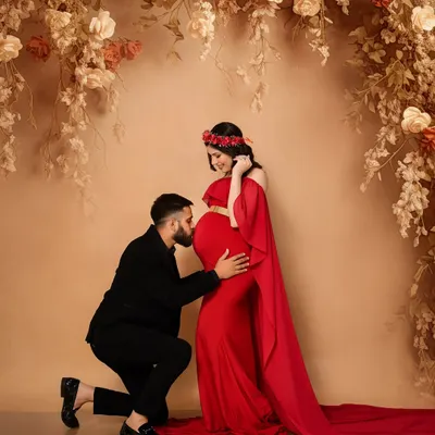 A classic and romantic maternity pose. The partner's gentle kiss on the baby bump against a floral-draped, fine art backdrop creates a timeless and passionate portrait. The vibrant red gown adds a touch of glamour to this special moment.