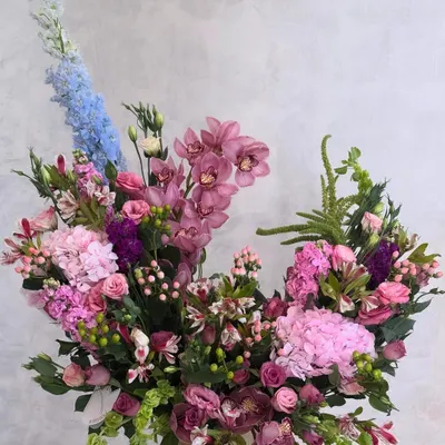 This is Wabi-Sabi in floral form. An asymmetrical arrangement that finds beauty in its imperfect, poetic shape, featuring a mix of pink hydrangeas, cymbidium orchids, and a single dramatic stem of blue delphinium.