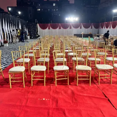 An outdoor wedding reception setup showing the guest seating arrangement. We used elegant gold chiavari chairs neatly arranged on a red carpet, with draped tents in the background, ready for an evening celebration.